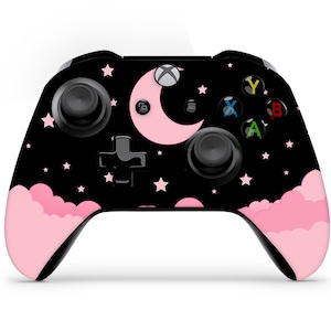 May include: A black Xbox controller with a pink and black design featuring a crescent moon and stars. The controller has pink buttons and a pink cloud design on the bottom.