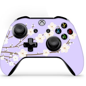 Könnte beinhalten: Ein lilafarbener Xbox-Controller mit einem floralen Design mit weißen Kirschblüten und braunen Zweigen. Der Controller hat schwarze Tasten und ein schwarzes Steuerkreuz.