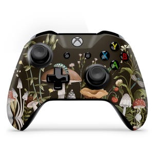 Könnte beinhalten: Ein schwarzer Xbox-Controller mit einem Design zum Thema Natur. Der Controller ist mit Illustrationen von Pilzen, Blumen und Blättern in Braun-, Grün- und Weißtönen verziert. Die Knöpfe sind in verschiedenen Farben gehalten.