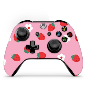 May include: A pink Xbox controller with a strawberry pattern. The controller features red strawberries and white flowers on a pink background. The controller has black joysticks, buttons, and a directional pad.