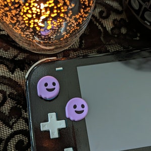 May include: A black Nintendo Switch console with two purple ghost-shaped button covers. The console is on a black lace surface.