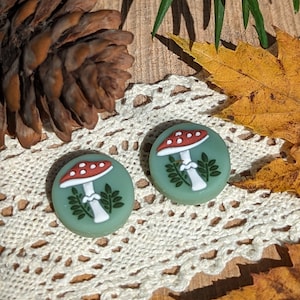 May include: Two round, green mushroom earrings with red and white mushroom designs. The earrings are set against a backdrop of a pinecone, lace, and fall foliage. The earrings are a whimsical accessory.