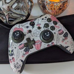 Puede incluir: Un mando Xbox blanco con un diseño de Halloween en rosa y negro. El mando presenta una variedad de símbolos e imágenes espeluznantes, incluyendo calabazas, calaveras, murciélagos y estrellas. El mando tiene dos botones negros con una cara sonriente en cada uno.