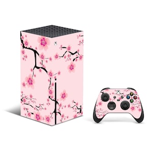 May include: A pink Xbox Series X console and controller with a cherry blossom design. The console is a rectangular prism with a black front panel and a black top. The controller is black with a white Xbox logo and white buttons. The design features pink cherry blossoms and black branches on a pink background.