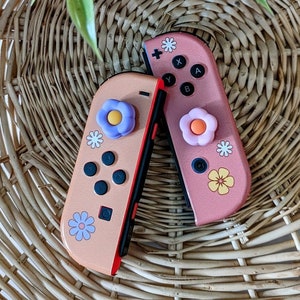 May include: Two pink and peach Nintendo Switch Joy-Con controllers with floral designs and colorful button covers. The controllers are on a woven wicker surface.