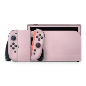 以下が含まれることがあります： ピンク色のNintendo Switch本体と、お揃いのJoy-Conコントローラー。コントローラーには黒いボタンとジョイスティックがあり、青と赤のアクセントが付いています。白い背景に置かれています。