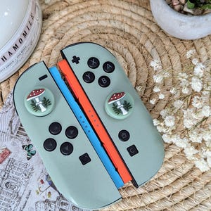 Op de afbeelding: Een mintgroene Nintendo Switch-controller met zwarte knoppen en paddenstoel-duimgrepen. De controller heeft oranje en blauwe rails. De afbeelding toont ook een kaars, een kleine plant en een ansichtkaart met Italiaanse tekst.