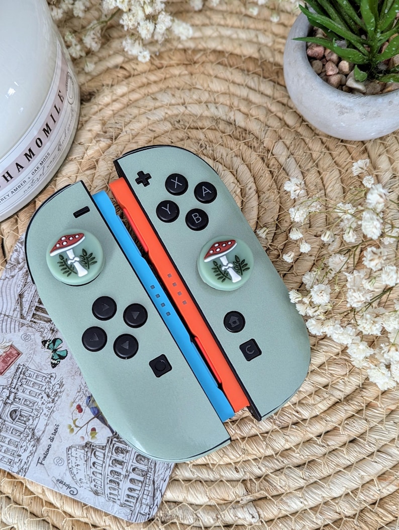 Op de afbeelding: Een mintgroene Nintendo Switch-controller met zwarte knoppen en paddenstoel-duimgrepen. De controller heeft oranje en blauwe rails. De afbeelding toont ook een kaars, een kleine plant en een ansichtkaart met Italiaanse tekst.