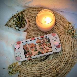 May include: A pink and white Nintendo Switch console with a peach design and a screen displaying a video game. The console is on a woven straw mat with a lit candle and a small potted plant.