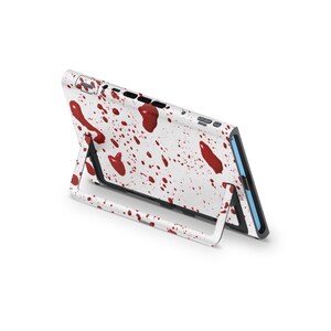 May include: White electronic device case with a red blood splatter design. The case is propped up on a white stand, showcasing the back and side of the device. The design features various sized red splatters.