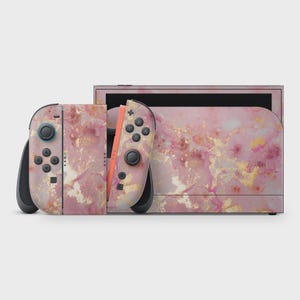 May include: A pink marble-patterned Nintendo Switch console and controllers. The controllers are detached, showing a coral-colored rail. The design features swirls of pink, gold, and white, creating a luxurious aesthetic. The console is set against a plain white background.