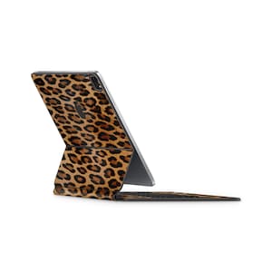 Puede incluir: Una tableta en una funda protectora con un estampado de leopardo en tonos marrones y negros. La funda está plegada en posición de soporte, dejando ver la parte posterior del dispositivo y la base del teclado integrada extendida horizontalmente.