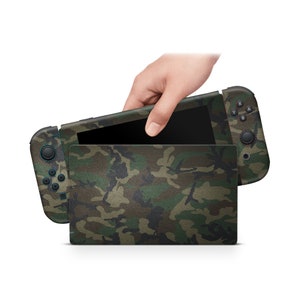 以下が含まれることがあります： 迷彩柄のNintendo Switch本体。黒い画面で、片手で上部を開けた状態。