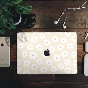 Könnte beinhalten: Ein Laptop mit einem weißen Gänseblümchenmuster auf dem Deckel. Der Laptop steht auf einem Holztisch mit einem weißen Telefon, weißen Ohrhörern und einer weißen Maus.