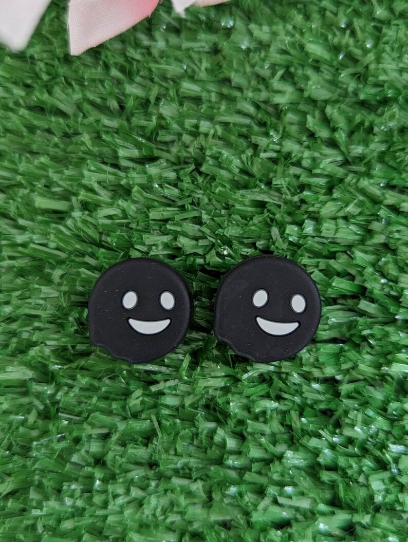 May include: Two black ghost-shaped phone charms with white smiling faces on a green artificial grass background.