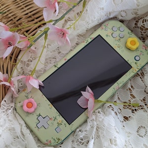 Może przedstawiać: Zielona konsola Nintendo Switch Lite z kwiatowym wzorem i różowymi i żółtymi osłonami przycisków. Konsola leży na białym koronkowej tkaninie.