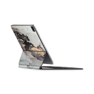 Puede incluir: Un iPad plateado con una funda de diseño de mármol blanco y dorado está plegado en forma de computadora portátil. El iPad está sobre una superficie blanca.