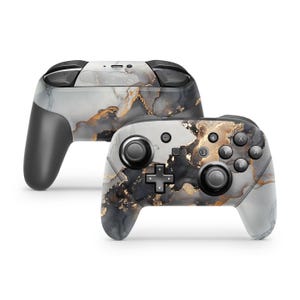 Puede incluir: Un mando de videojuegos inalámbrico con un patrón de mármol en tonos negro, blanco y gris con detalles en oro metálico. El mando incluye una cruceta, dos joysticks analógicos y botones etiquetados con las letras A, B, X e Y.