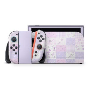 May include: A lavender and white Nintendo Switch console with matching Joy-Con controllers. The console and controllers feature a patchwork floral design in shades of purple, pink, and white. The controllers have black buttons and joysticks.