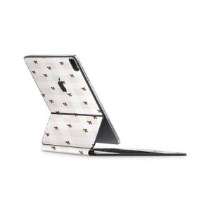 以下が含まれることがあります： 白とベージュのチェック柄に小さな茶色のリボンが施されたApple iPad Pro。 iPadは開いており、画面とキーボードが表示されています。 Appleのロゴがデバイスの背面に表示されています。