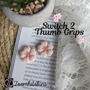 May include: Two pink flower-shaped thumb grips for a Switch 2 controller. The grips are light pink with a darker pink centre. The text "Switch 2 Thumb Grips" is displayed on the background.