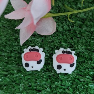 May include: Two white cow-shaped silicone charms with black spots and pink noses. The charms are on a green artificial grass background.