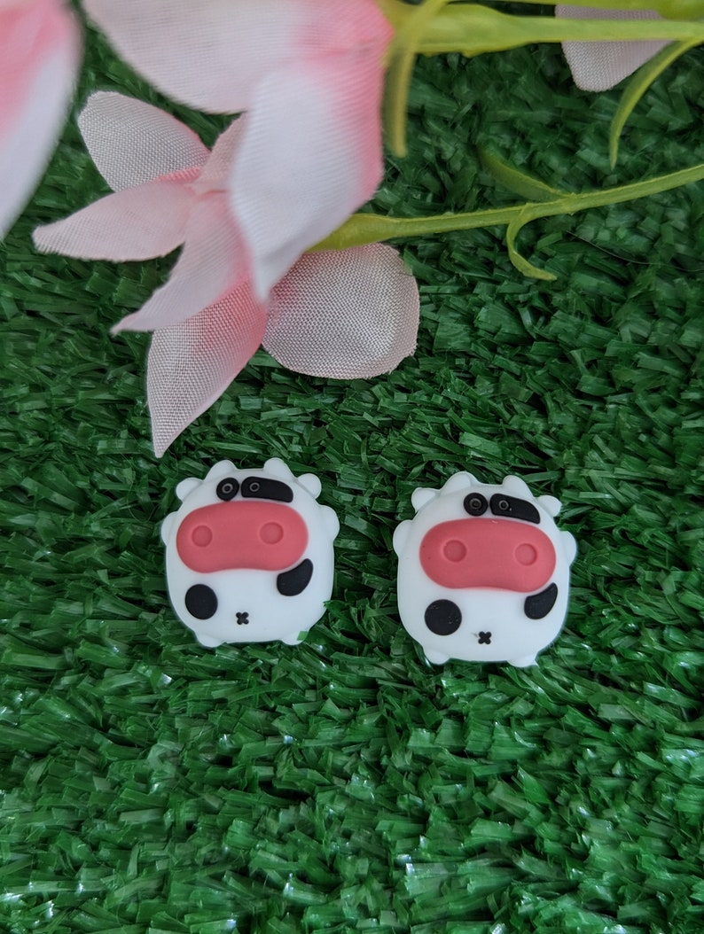 May include: Two white cow-shaped silicone charms with black spots and pink noses. The charms are on a green artificial grass background.