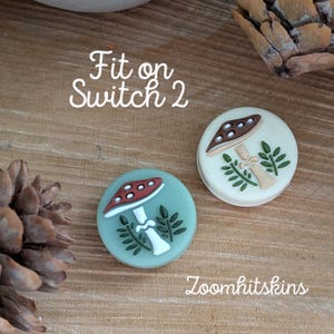 May include: Two round decorative accessories with mushroom designs. One is light blue with a red-and-white mushroom, the other is white with a brown-and-white mushroom. The text "Fit on Switch 2" and "Zoomhitskins" are also visible.