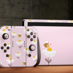 Pode incluir: Consola Nintendo Switch rosa e controladores Joy-Con com um design de margaridas e abelhas. A consola e os controladores são decorados com um padrão de margaridas brancas e abelhas amarelas sobre um fundo rosa.