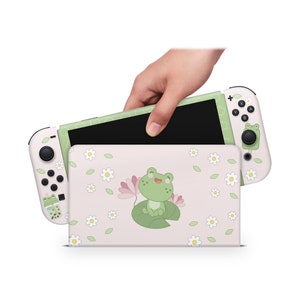 Puede incluir: Una piel para consola Nintendo Switch de color rosa y verde con un diseño de rana adorable. La piel presenta una rana sentada en un nenúfar con flores rosas y hojas verdes. La consola se muestra cerrada con la pantalla mirando hacia la cámara.