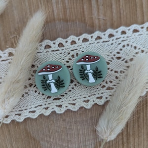 Op de afbeelding: Twee ronde, saliegroene oorbellen met een paddenstoelontwerp. De paddenstoelen hebben rood-witte doppen, witte stelen en zwarte bladeren. De oorbellen worden gepresenteerd op een wit kanten lint en een houten oppervlak.