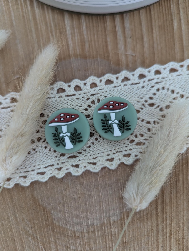 Op de afbeelding: Twee ronde, saliegroene oorbellen met een paddenstoelontwerp. De paddenstoelen hebben rood-witte doppen, witte stelen en zwarte bladeren. De oorbellen worden gepresenteerd op een wit kanten lint en een houten oppervlak.