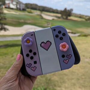 May include: A purple and white Nintendo Switch console with a pink heart and flower designs. The console has a black grip and pink buttons.