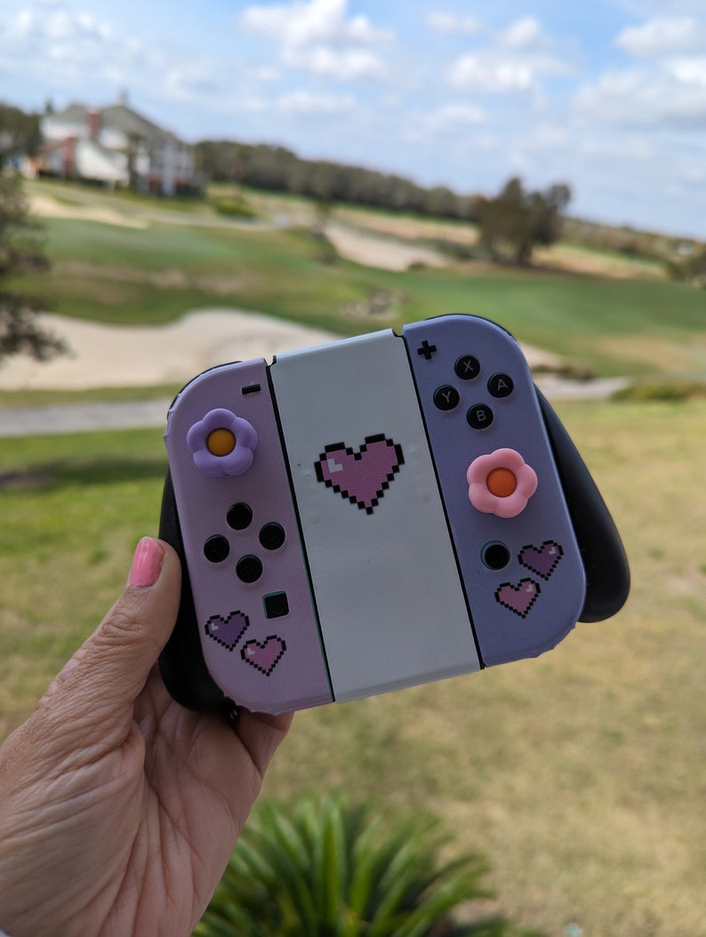 May include: A purple and white Nintendo Switch console with a pink heart and flower designs. The console has a black grip and pink buttons.