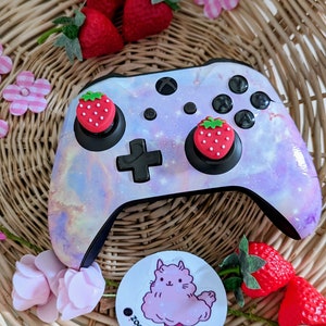 Può includere: Un controller Xbox bianco e viola a tema galassia con due cappucci a forma di fragola. Il controller è appoggiato su un cesto di vimini con fiori rosa e un adesivo di un alpaca rosa che mangia zucchero filato.