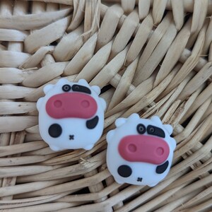 May include: Two white cow-shaped silicone charms with black spots and pink noses. The charms are sitting on a woven wicker surface.