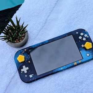 May include: A blue and gold Nintendo Switch Lite console with a galaxy design. The console has yellow star-shaped buttons.