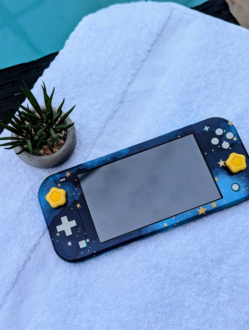 May include: A blue and gold Nintendo Switch Lite console with a galaxy design. The console has yellow star-shaped buttons.