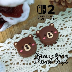 May include: Two brown bear-shaped thumb grips for a Nintendo Switch controller. Each grip features a bear face with pink cheeks and a light brown muzzle. The Nintendo Switch logo is visible in the background.