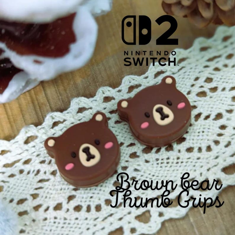 May include: Two brown bear-shaped thumb grips for a Nintendo Switch controller. Each grip features a bear face with pink cheeks and a light brown muzzle. The Nintendo Switch logo is visible in the background.