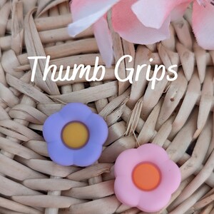 May include: Two flower-shaped thumb grips, one purple with a yellow center and one pink with an orange center. The grips are on a woven wicker surface. "Thumb Grips" is written in white text on the surface.
