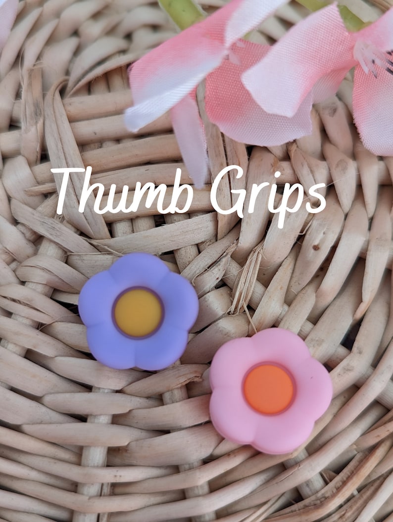 May include: Two flower-shaped thumb grips, one purple with a yellow center and one pink with an orange center. The grips are on a woven wicker surface. "Thumb Grips" is written in white text on the surface.