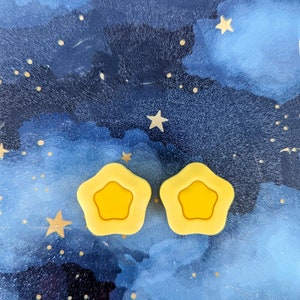 May include: Two yellow star-shaped hair clips against a blue and white watercolor background with gold glitter stars.