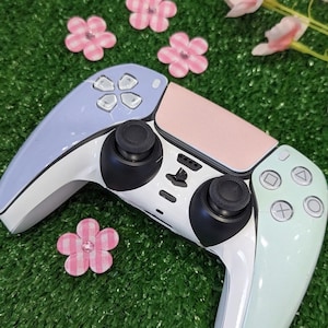 Peut inclure: Une manette de jeu vidéo personnalisée avec des accents de couleurs pastel. La manette présente un dessus bleu clair, un centre rose et un côté droit vert menthe. Les boutons et les joysticks sont noirs, et la base est blanche.