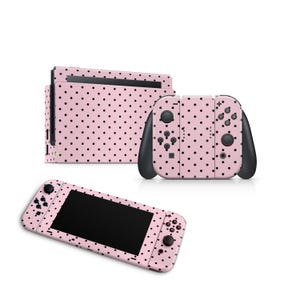 Könnte beinhalten: Eine rosa Nintendo Switch-Konsole und -Controller mit einem schwarzen Polka-Dot-Muster. Das Set enthält die Hauptkonsole, zwei Joy-Con-Controller und die Dockingstation. Die Joy-Con-Controller haben schwarze Akzente.