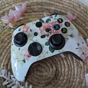 May include: A white Xbox controller with a floral pattern featuring pink roses and green leaves. The controller has black buttons and a black D-pad.
