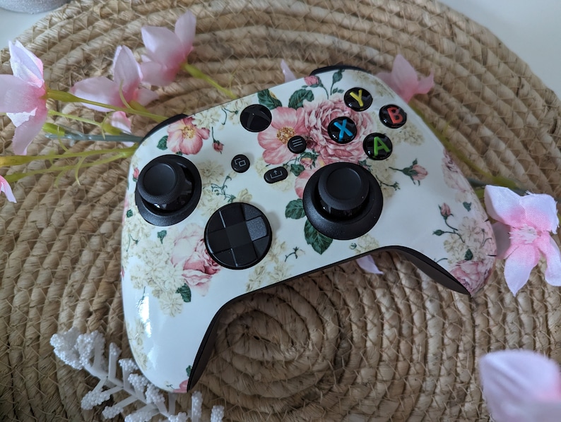 May include: A white Xbox controller with a floral pattern featuring pink roses and green leaves. The controller has black buttons and a black D-pad.