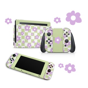 Pode incluir: Um console Nintendo Switch xadrez verde e branco com detalhes de flores roxas. O console é mostrado em três partes: o console principal, o controlador Joy-Con esquerdo e o controlador Joy-Con direito.