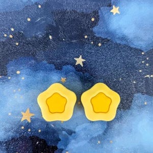 May include: Two yellow star-shaped hair clips against a blue watercolor background with white stars.