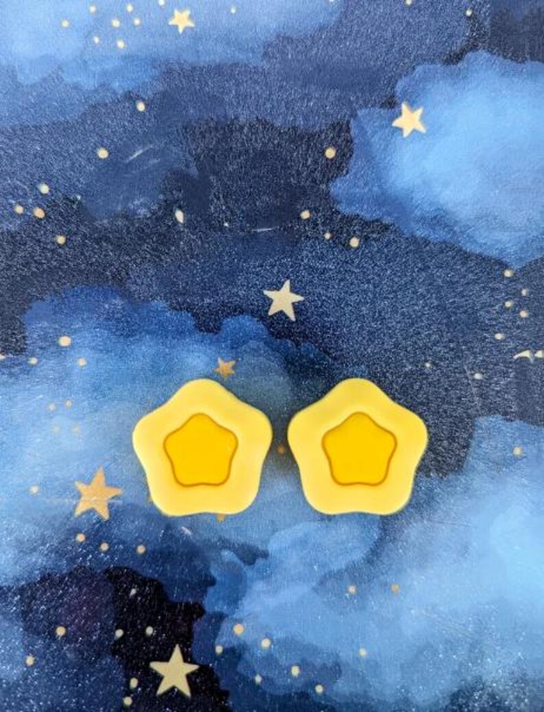 May include: Two yellow star-shaped hair clips against a blue watercolor background with white stars.